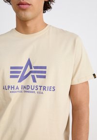 Beige bomulds t-shirt med et lilla grafisk logo med vinger og teksten "ALPHA INDUSTRIES" nedenunder, rund halsudskæring og korte ærmer.