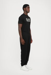 Camiseta de algodón negra con logotipo impreso, combinada con pantalones de chándal negros y zapatillas blancas. Diseño minimalista, corte estándar y cinturilla elástica.