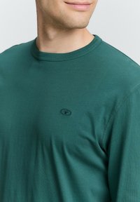 Grünes Baumwoll-T-Shirt mit rundem Ausschnitt, das ein kleines schwarzes Logo auf der Brust und kontrastierende Nähte entlang der Kanten aufweist.
