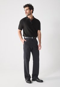 Schwarzes Polohemd mit Knöpfen, kombiniert mit dunklen Hosen und einem schwarzen Ledergürtel. Das Modell trägt schwarze Schuhe und steht vor einem weißen Hintergrund.