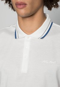 Camisola polo branca feita de tecido texturizado. Apresenta um colarinho clássico com riscas azuis e cinzas e uma pala com três botões. Logótipo no peito.