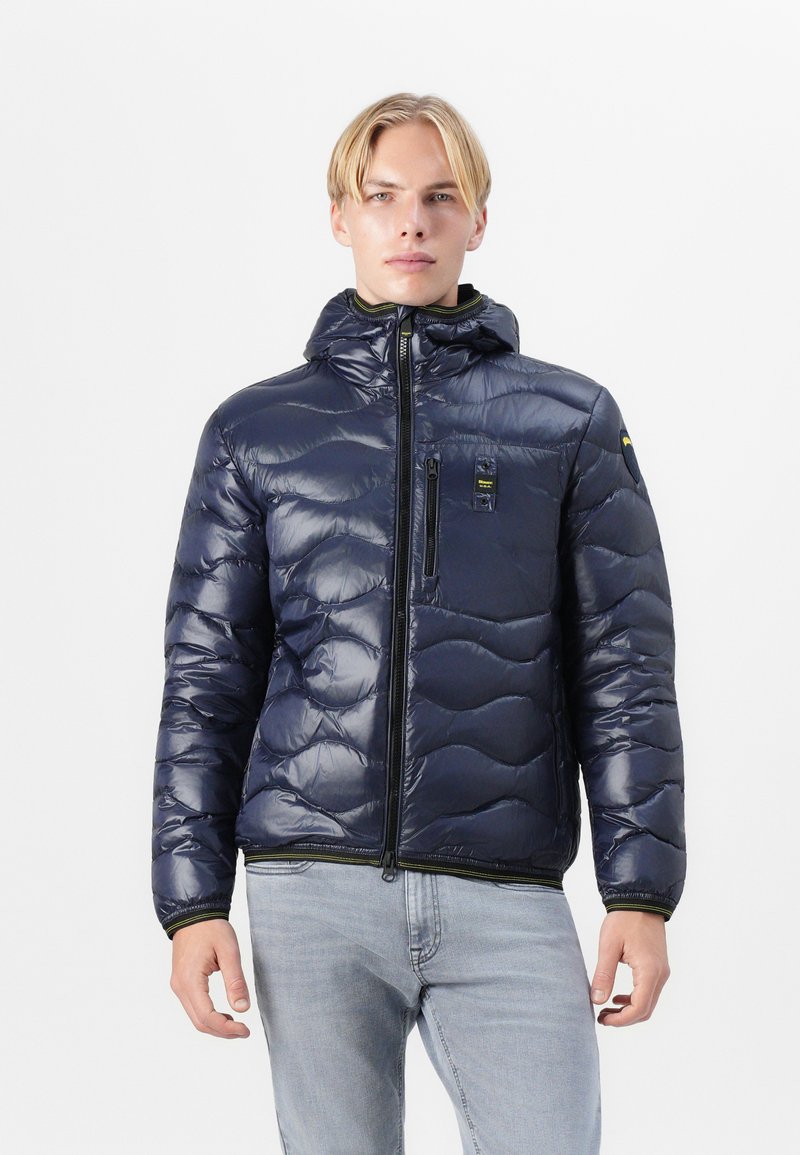 Doudoune marine avec capuche, design matelassé, poches zippées et légère brillance. Accentuée de détails en vert lime et de marquages.