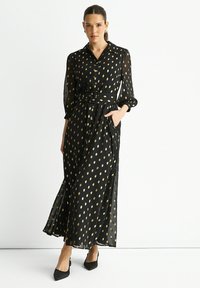Black maxi dress with gold leaf patterns, long sheer sleeves, a belted waist, and side pockets. Styled with pointed black shoes.