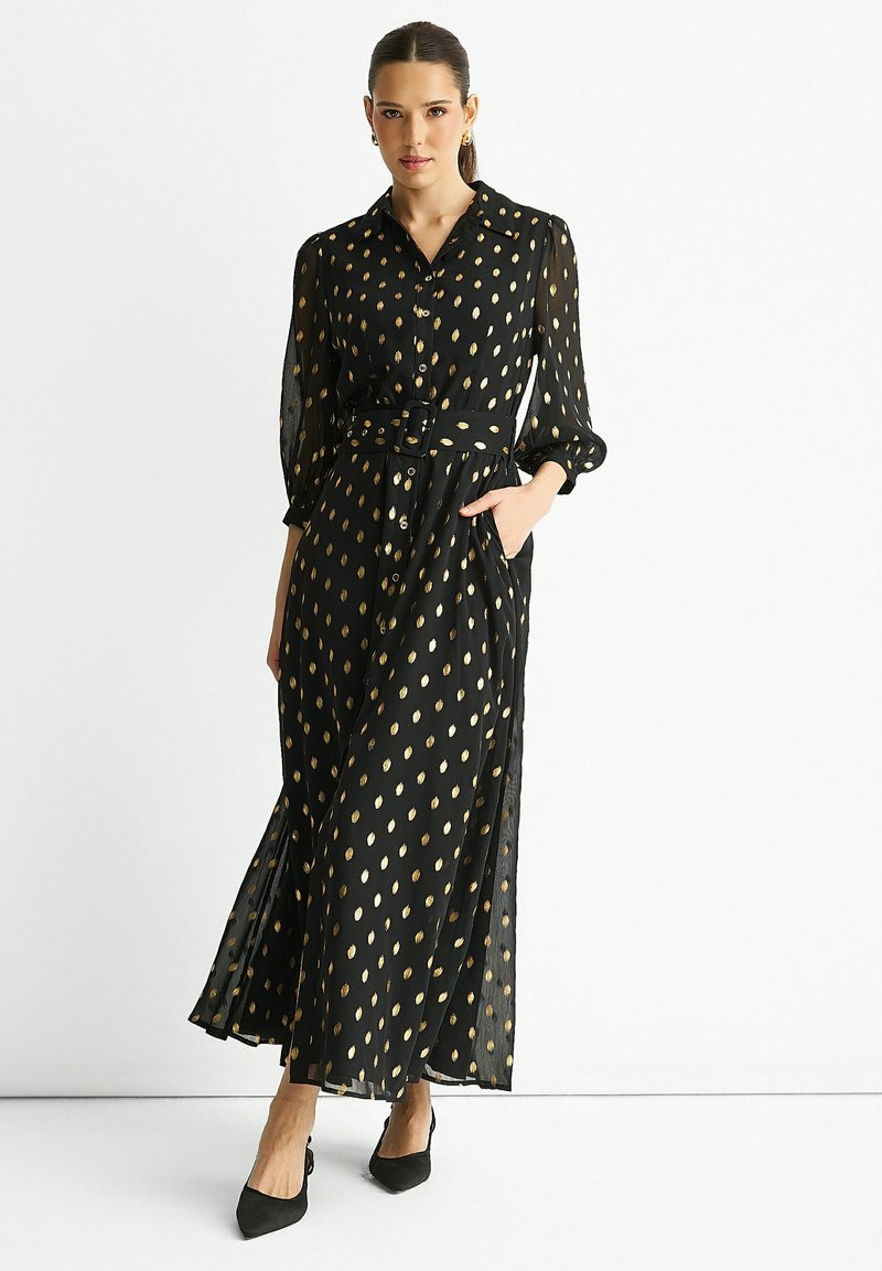 Black maxi dress with gold leaf patterns, long sheer sleeves, a belted waist, and side pockets. Styled with pointed black shoes.