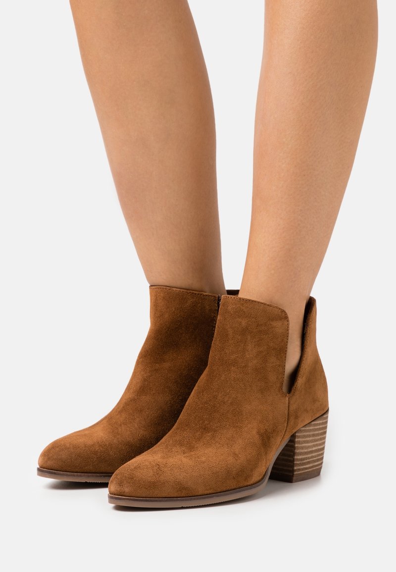 Botas de tobillo de ante marrón con un tacón bajo y apilado, punta afilada y recortes laterales. El material tiene una textura suave con un acabado mate.