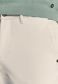 Witte broeken met een gladde textuur, voorzien van een knoopsluiting, zijzakken en doorgestikte details. Gecombineerd met een lichtgroene trui.
