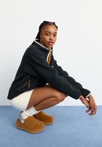 Μαύρο φλις πουλόβερ με καφέ γιακά, τσέπη με φερμουάρ στο πλάι, λευκή γαζιά. Ανοιχτό μπεζ φούστα, υφασμάτινα καφέ ξύλινα παπούτσια με γκρι λεπτομέρειες, λευκές κάλτσες.