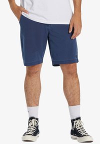 Herren Shorts in Marineblau aus Baumwolle, mit seitlichen Taschen und geradem Schnitt, der kurz über dem Knie endet. Getragen mit schwarzen hohen Sneakers.