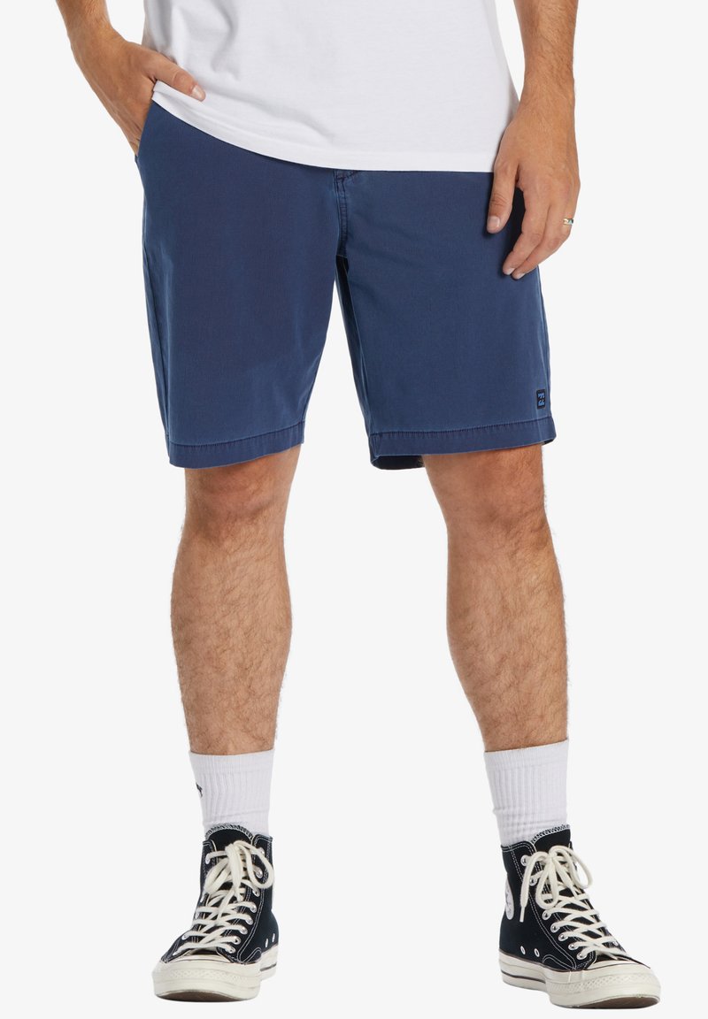 Herren Shorts in Marineblau aus Baumwolle, mit seitlichen Taschen und geradem Schnitt, der kurz über dem Knie endet. Getragen mit schwarzen hohen Sneakers.
