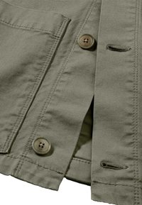 Olijfgroen jack met twee voorkanten zakken, grijze knopen en diagonale stiksels. De stof heeft een gestructureerd oppervlak en zichtbare naden.