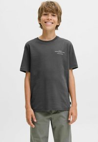 Smiling boy wearing a dark gray t-shirt with text on the front, standing against a plain white background, hands relaxed at his sides.