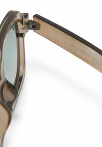Close-up van het scharnier en de arm van een bruin, doorschijnend zonnebrilmodel met "JACK & JONES" branding aan de binnenzijde van de arm.