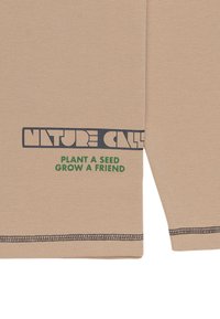 Beige strukturerad tyg med "NATURE CALLS" tryckt i mörkblått och "PLANT A SEED GROW A FRIEND" i grönt. Sömmen har mörka sömnadsdetaljer.