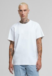 Witte katoenen t-shirt met korte mouwen, relaxte pasvorm en ronde hals, gedragen met lichtblauwe jeans. Bevat zichtbare tatoeages op de armen.