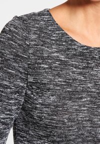 Grå och svart strukturerad tröja med rund halsringning, med ett subtilt melerat mönster och mjukt, elastiskt tyg. Närbild.