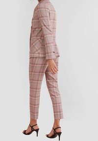 Costume à carreaux roses comprenant un blazer cintré et un pantalon sur mesure. Confectionné dans un tissu texturé avec une finition lisse. Accessoirisé avec des talons noirs.