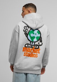Grå hettegenser med et fargerikt tegneserie-jordklodegrafikk på baksiden, med "market studios" tekst i oransje under. Materiale av bomullsblanding.