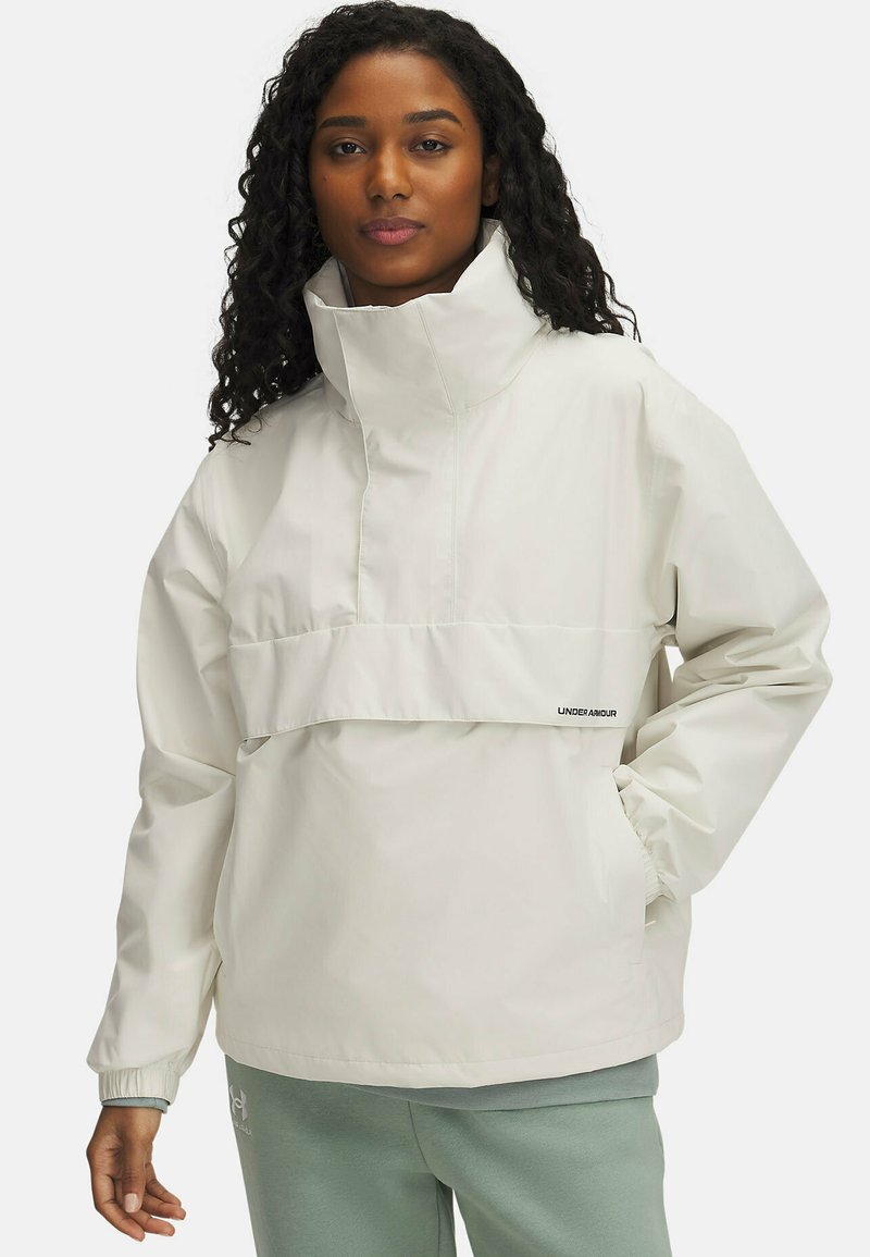 Lightweight cream rain jacket with high collar, front pocket, and elastic cuffs. Under Armour logo near the hem. Worn over gray sweatpants.