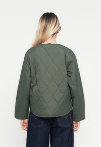 Personne avec de longs cheveux blonds portant une veste matelassée olive et un jean bleu foncé, se tenant face à un fond blanc.