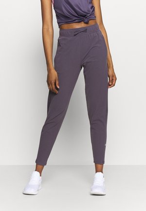 Femme portant un pantalon de jogging taille haute, ajusté, violet foncé, et des baskets blanches, debout devant un fond clair uni.