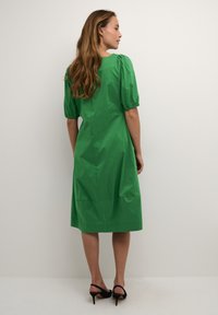 Rochie verde, până la genunchi, dintr-un material neted, cu mâneci bufante, decolteu rotund și talie strânsă, purtată cu sandale negre cu toc.