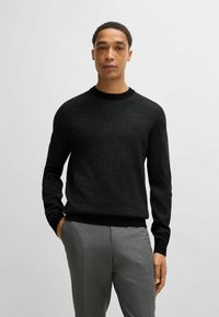 Maglione nero lavorato a maglia con un motivo testurizzato, scollatura tonda e polsini e bordo a costine, abbinato a pantaloni grigi.