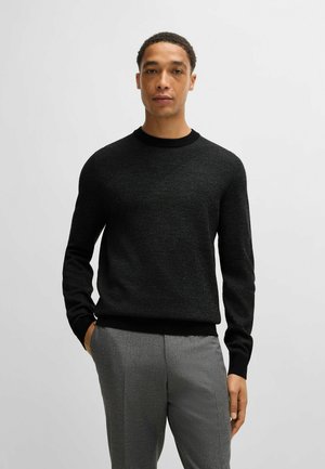 Maglione nero lavorato a maglia con un motivo testurizzato, scollatura tonda e polsini e bordo a costine, abbinato a pantaloni grigi.