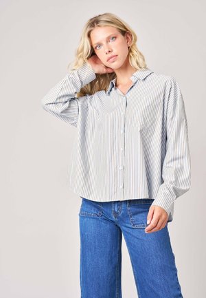 Jeune femme aux longs cheveux blonds porte une chemise large à rayures boutonnée et un jean bleu, posant avec une main derrière la tête.