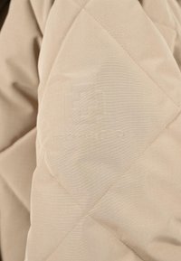 Béžová prošívaná bunda s jemným vodorovným pruhem. Na levém rukávu se nachází debossované logo.
