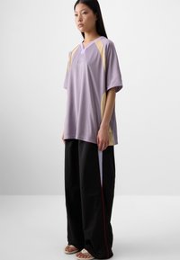 Une jeune femme se tient devant un fond uni, portant un top oversized lavande avec des panneaux en maille et un pantalon noir à jambes larges.