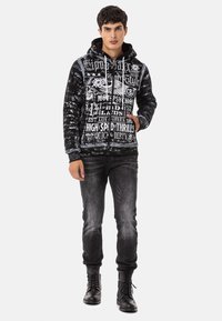 Schwarz-weiße grafische Kapuzenjacke mit Text und Totenkopfdesign, kombiniert mit verwaschenen schwarzen Jeans und schwarzen Schnürstiefeln, die einen lässigen Stil präsentieren.