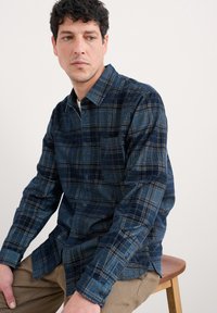 Man in een blauw geruit overhemd met knopen en beige broek, zittend op een houten kruk, kijkend naar links tegen een witte achtergrond.