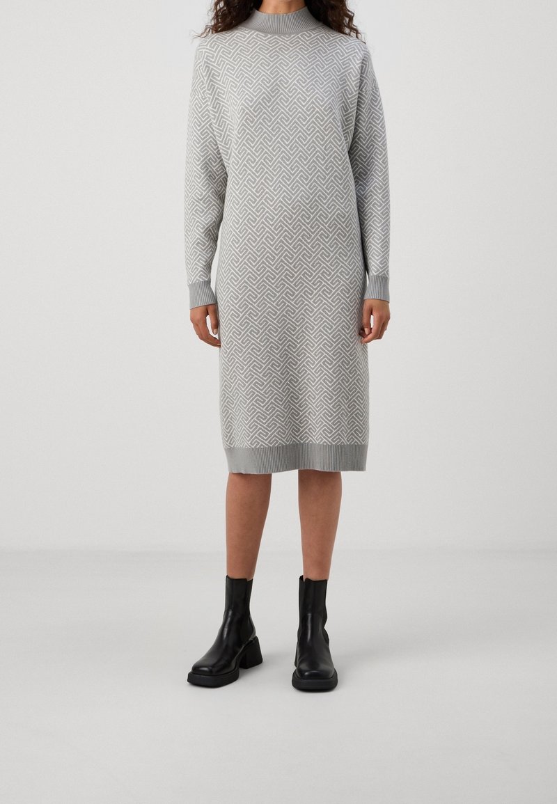 Femme portant une robe en maille grise à motifs géométriques et manches longues, associée à des bottines noires, debout sur un sol gris clair.
