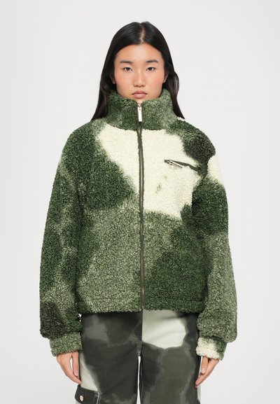 Chaqueta de forro polar en verde y crema, con un alto cuello, cierre frontal y un bolsillo en el pecho con cremallera que tiene una apariencia texturizada.