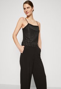 Débardeur noir avec des fines bretelles, orné de clous en argent disposés en motif de grille. Associé à un pantalon noir taille haute, texture lisse.