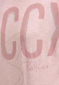 Hellrosa T-Shirt mit großem, zartrosa Schriftzug und silbernen Strass-Akzenten. Der Stoff hat ein glattes, leicht strukturiertes Erscheinungsbild.