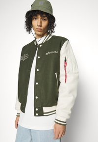 Giacca varsity verde e crema con chiusura a bottoni, polsini a costine, loghi ricamati e un'etichetta rossa sul maniche.
