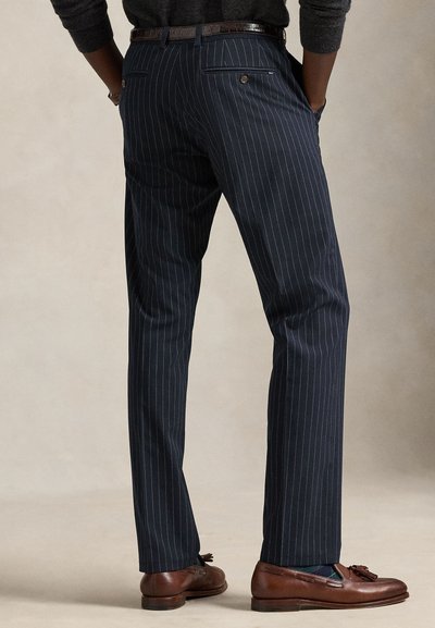 Man wearing navy blue pinstripe trousers, brown belt, dark sweater, and brown leather tassel loafers standing with hands in pockets.