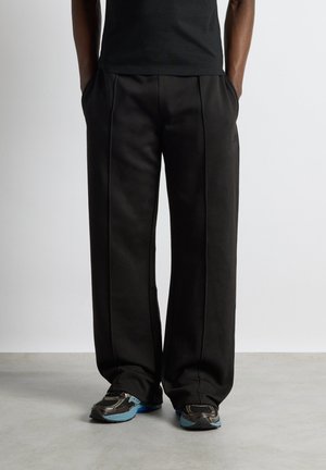 Persoană purtând pantaloni negri largi și adidași negri cu tălpi albastre, stând pe un podea gri deschis, în fața unui perete alb simplu.