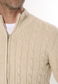Beige gestrickter Zip-Up-Pullover mit einem gerippten Kragen und Zopfmuster. Verfügt über einen Front-Reißverschluss und strukturierter Stoff.