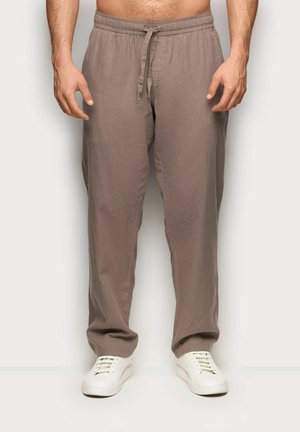 Mężczyzna w luźnych brązowych spodniach ze sznurkiem i białych trampkach, stojący na neutralnym tle.