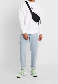 Uomo che indossa una felpa bianca, jeans leggermente blu arrotolati, sneakers verde neon e borsa a tracolla nera su sfondo bianco.