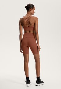 Športni modrček v rjavo-rdeči barvi z naramnicami v obliki križa in ujemajočimi visokimi kratkimi hlačami. Narejeno iz gladkega, raztegljivega materiala.