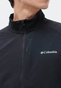 Άνδρας που φοράει μαύρο μπουφάν Columbia με φερμουάρ και ψηλό γιακά, που δείχνει μέρος από το στήθος και τον λαιμό στρεφόμενα προς τα πλάγια.
