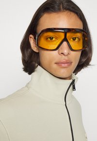Occhiali da sole con grandi lenti gialle e montatura nera, indossati da una persona con i capelli lunghi, abbinati a una felpa beige con zip.