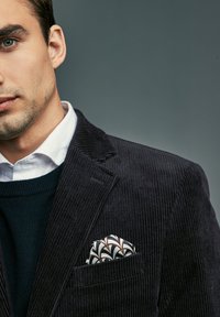 Junger Mann mit weißem Hemd, marineblauem Pullover und dunklem Cordblazer mit gemustertem Einstecktuch vor grauem Hintergrund.
