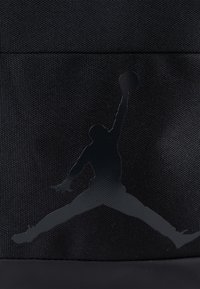 Mochila negra con un logotipo de Jordan en relieve y brillo en una pose de salto, hecha de un tejido texturizado con un panel inferior liso.