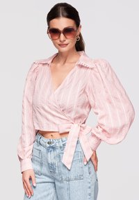 Femme portant des lunettes de soleil carrées surdimensionnées, un chemisier rose portefeuille avec des manches bouffantes et un jean taille haute bleu clair, main sur la hanche.