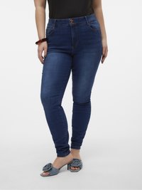High-waisted, skinny-fit jeans in dark blue denim with five pockets and contrasting stitching; paired with grey heeled sandals featuring a floral accent.
