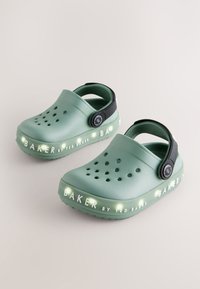 Groene rubberen klompen met een zwarte band, voorzien van ventilatiegaten en verlichte tekst die zegt "BAKER by TED BAKER" langs de zool.